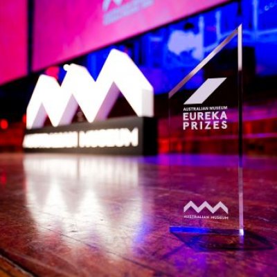 A photo of a stage, lit up with pink and blue lights, and a glass trophy in the foreground saying Eureka Prizes.